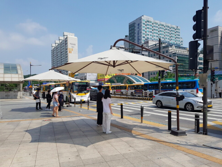 여름철 횡단보도 앞 쉼터를 만들어주는 파라솔형 그늘막 밑에 서 있는 사람들 이미지